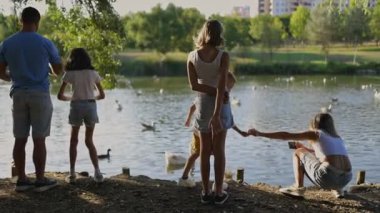 Aile, gölün kenarında güneşli bir öğleden sonranın tadını çıkarıyor. Bir kadın anı yakalarken ördekleri besliyor.