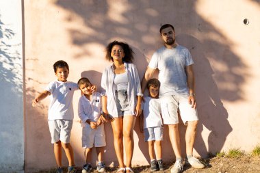 Pembe bir duvara karşı poz verip güneşli bir günün tadını çıkaran çok ırklı mutlu bir aile.