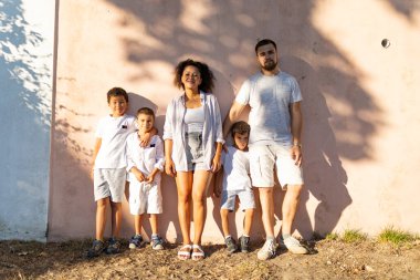 Pembe bir duvara karşı poz verip güneşli bir günün tadını çıkaran çok ırklı mutlu bir aile.