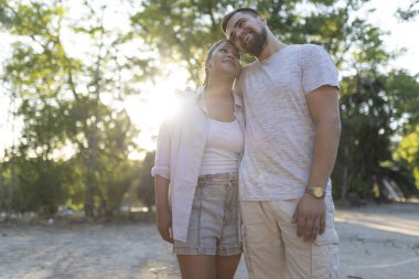 Parkta kucaklaşan genç çift gün batımında romantik bir anın tadını çıkarıyor.
