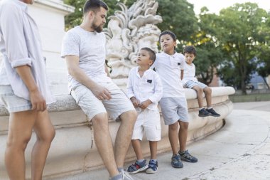 Baba ve üç oğlu yaz tatili boyunca şehir parkındaki çeşmede dinlenirken anneleri yanlarında duruyor.