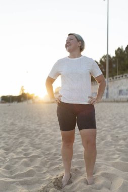 Kıvrımlı sporcu kadın gün batımında sahilde huzur ve güven dolu bir anın tadını çıkarıyor, vücut pozitifliğini ve aktif bir yaşam tarzını destekliyor.