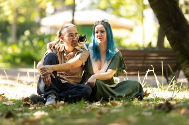 Parkta çimenlerde oturan genç bir çift. Yavruları erkeğin omzunda güneşli bir günün tadını çıkarıyorlar.