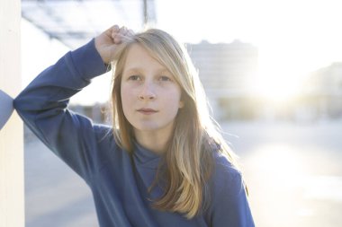 Uzun sarı saçlı, kendine güvenen genç bir kızın portresi bulanık bir şehir ortamında dikilirken saçına dokunuyor.