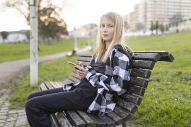 Şehir parkında bankta otururken cep telefonu uygulaması kullanan sarışın bir kız.