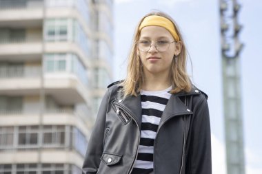Gözlüklü ve deri ceketli şık bir genç kız şehir zemininde poz veriyor.