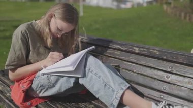 Parktaki bankta otururken not defterine yazan konsantre öğrenci kız.