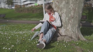 Parkta kitap okumaktan keyif alan huzurlu bir öğrenci güneşli bir günde papatya topluyor.