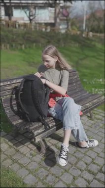 Genç öğrenci sırt çantasından bir kitap alıyor ve parkta bir bankta otururken onu okuyor.