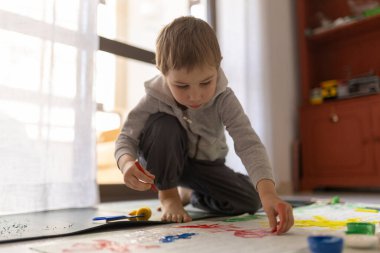 Resim yapmaya odaklanan, parmak ve fırça kullanan, sanatsal ifade ve çocukluk gelişimi gösteren genç bir çocuk.