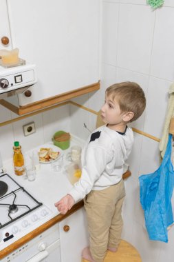 Genç çocuk bir taburede duruyor, mutfak tezgahında bırakılan yiyeceklere uzanıyor.