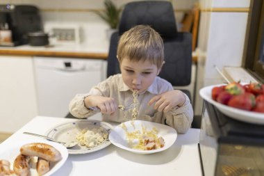 Sarışın çocuk, mutfaktaki makarnanın ve sosisin tadını çıkarıyor. Yanında da çilek var.