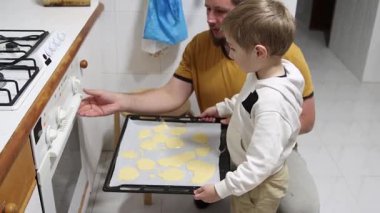 Baba küçük oğluna kurabiye pişirmesi için yardım ediyor, tepsiyi fırına koyuyor.