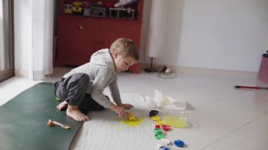 Genç çocuk yaratıcı oyun zamanını seviyor, yere saçılmış kağıtta sarı boya ile resim yapıyor.