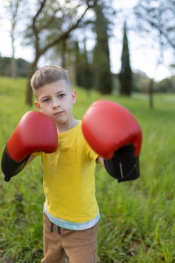 Çocuk parkta boks yapıyor, kırmızı boks eldiveni ve sarı tişört giyiyor.