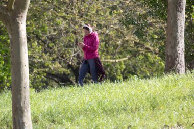 Yeşil bir parkta rahatça dolaşırken kulaklıkla müzik dinleyen bir kadın.