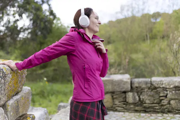 Yürüyüşçü doğada molada müziğin keyfini çıkarıyor, pembe ceket ve kulaklık giyiyor.