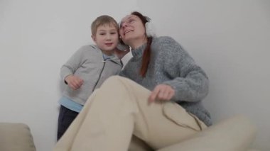 Mutlu anne ve oğul birlikte müzik dinliyorlar, kulaklık takıyorlar ve beyaz bir arka plana karşı kanepede dans ediyorlar.