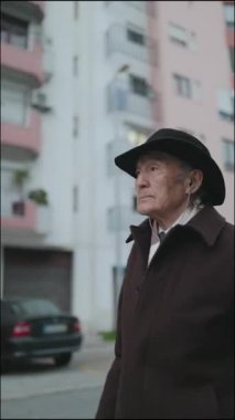 Şapka ve kulaklık takan düşünceli yaşlı vatandaş kentsel çevrede çevreyi gözlemliyor