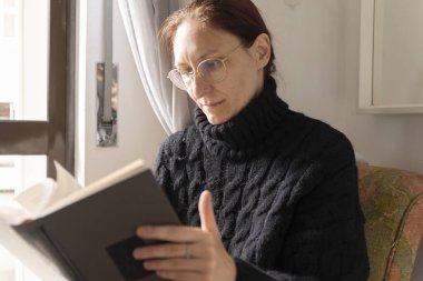 Orta yaşlı, gözlüklü bir kadın kitap okumaktan zevk alırken pencerenin kenarında rahat bir şekilde oturuyor.