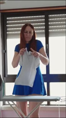 Mavi elbiseli kızıl kadın ütü masasında beyaz tişörtü inceliyor.