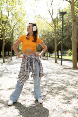 Güneşli bir günde, şehir parkında müzik dinleyen kulaklık takan mutlu genç bir kadın.