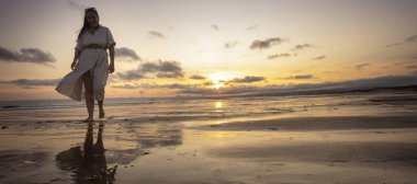 Gün batımında ıslak sahilde yürüyen bir kadın, suda yansıyan bir yansımayla.