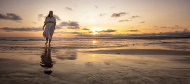 Islak kumda yürüyen manken plajdaki altın gün batımını yansıtıyor.