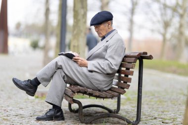 Park bankında otururken deftere yazı yazan kıdemli yazar, huzurlu bir ortamın tadını çıkarıyor.