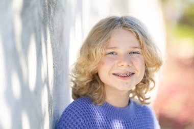 Sakallı sarışın kız gülümsüyor, gölgeli duvara yaslanıyor.