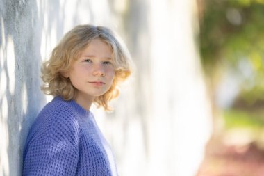 Açık renkli bir duvara yaslanmış, benekli gölgeleri olan, mor bir kazak giyen genç bir kadın.