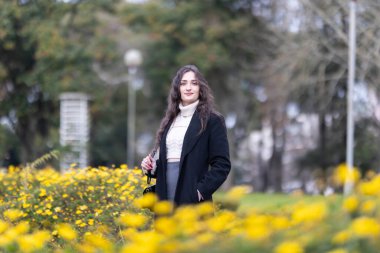 Açık havada poz veren genç bir kadın halka açık bir parkta canlı sarı çiçeklerle çevrili.