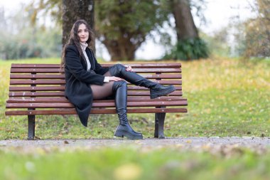 Dışarıdaki ahşap bir bankta oturan şık siyah çizmeler ve paltolar giyen gülümseyen kadın.