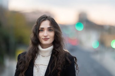 Bulanık bir şehrin arka planında dururken gülümseyen genç bir kadın.