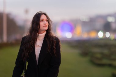 Parkta duran genç bir kadın, özenle arka plandaki şehir ışıklarına bakıyor.