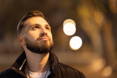 Sakallı genç adam yukarı bakıyor, şehir gece ortamında geleceği düşünüyor.
