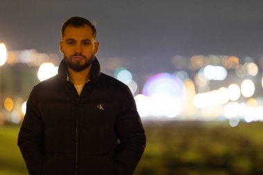 Siyah balon ceketli sakallı bir adam. Arkasında bulanık şehir ışıkları olan kameraya bakıyor.