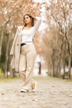 Asfalt bir yolda duran genç bir kadın, parktaki sonbahar atmosferinin tadını çıkarıyor.