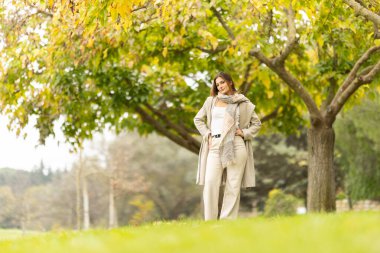 Genç bir kadın parkta duruyor, modern bir ceket ve eşarp giyiyor, kameraya bakıyor.