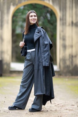 Paltosu ve geniş paçalı pantolonuyla şık bir kıyafetin içinde gülümseyen genç bir kadın.