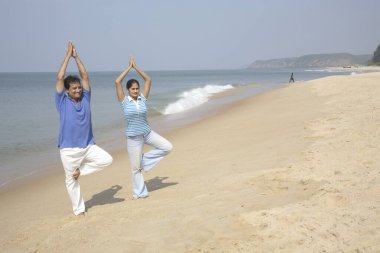 Güney Asyalı genç adam ve kadın yogayı tek ayak üzerinde yapıyor ve deniz kıyısında, Shiroda, Dist Sindhudurga, Maharashtra, Hindistan 'da avuç içlerini birleştiriyor.  