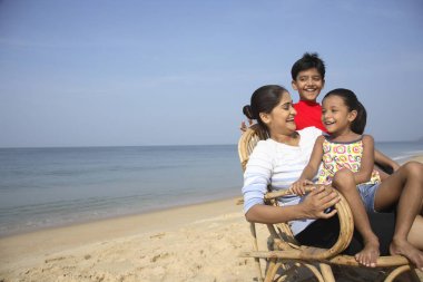Güney Asyalı Hintli kız baston sandalyede annesinin kucağında ve oğlu deniz kıyısında ayakta duruyor.