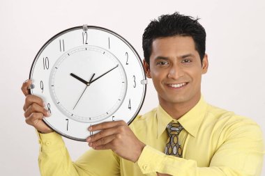 İş adamı duvar saatini iki elinde tutuyor. Sağ tarafının yanında. 