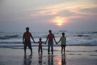 Güney Asyalı ebeveynler ve çocuklar deniz kıyısında altın gün batımını seyrediyorlar, Shiroda, Dist Sindhudurga, Maharashtra, Hindistan