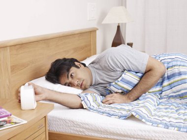 Young man laying on bed hand stopping ringing alarm clock  clipart