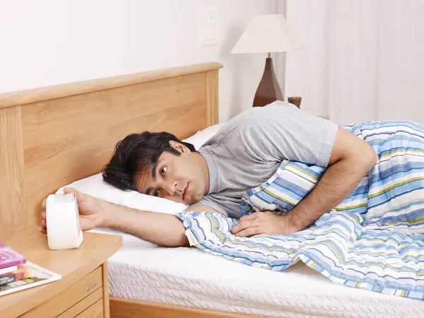 stock image Young man laying on bed hand stopping ringing alarm clock 