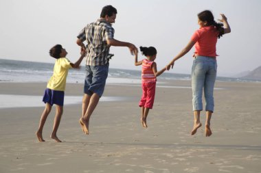 Güney Asyalı ebeveynler ve çocuklar deniz kıyısında el ele zıplıyorlar, Shiroda, Dist Sindhudurga, Maharashtra, Hindistan 