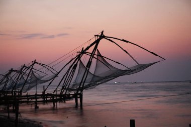 Çin balıkçı ağları akşam geç saatlerde, Hindistan, Kerala, Ernakulum Bölgesi, Fort Kochi 'deki uzak gemilerde kırmızımsı renkli gökyüzü ışıkları ile kırmızıya boyandı.  