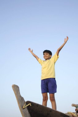 Güney Asyalı çocuk ahşap bir teknede önü açık duruyor. İki elini de sahile doğru açıyor.