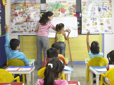 Güney Asyalı Hintli bir kız tahtaya yazı yazıyor ve öğretmen anaokulunda ona yardım ediyor.  